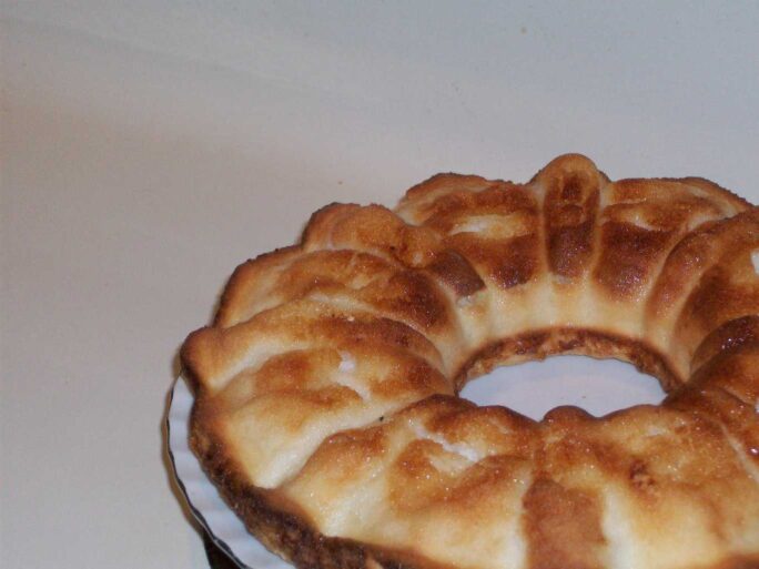 A ring-shaped baked cake with a golden-brown top sits on a plate against a white background.