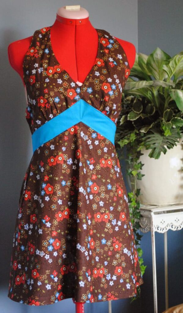 The red mannequin displays a sleeveless brown dress with colorful floral patterns and a blue waistband, set against a backdrop featuring a potted plant—perfect for Picture Tuesday.