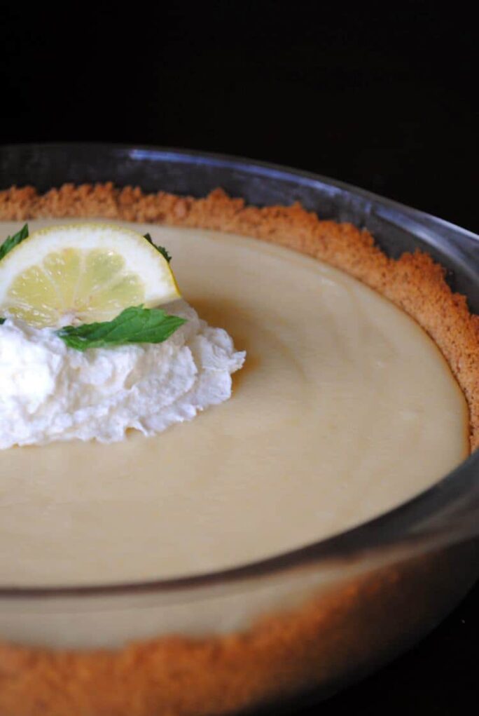 A close-up of an easy lemon pie with a graham cracker crust, topped with whipped cream, a lemon slice, and a mint leaf.