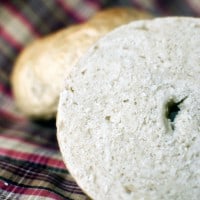 A sliced, bakery-fresh bagel rests on a plaid cloth with a whole bagel gleaming in the background.