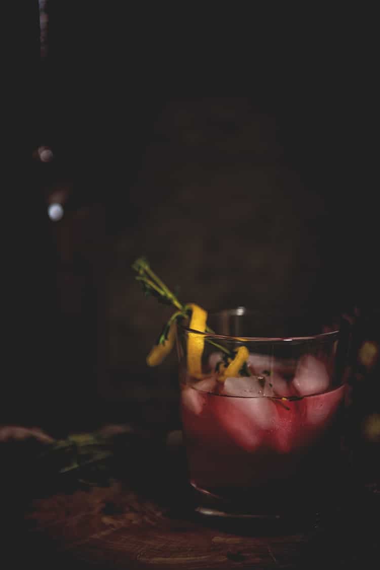 Close up of highball glass filled with ice and huckleberry vodka cocktail, garnished with lemon twist and thyme sprigs. 