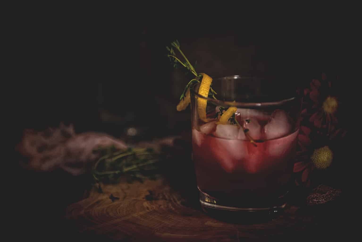 Dark moody photo of a highball glass with purple huckleberry vodka cocktail garnished with lemon twist wrapped around thyme sprigs. 