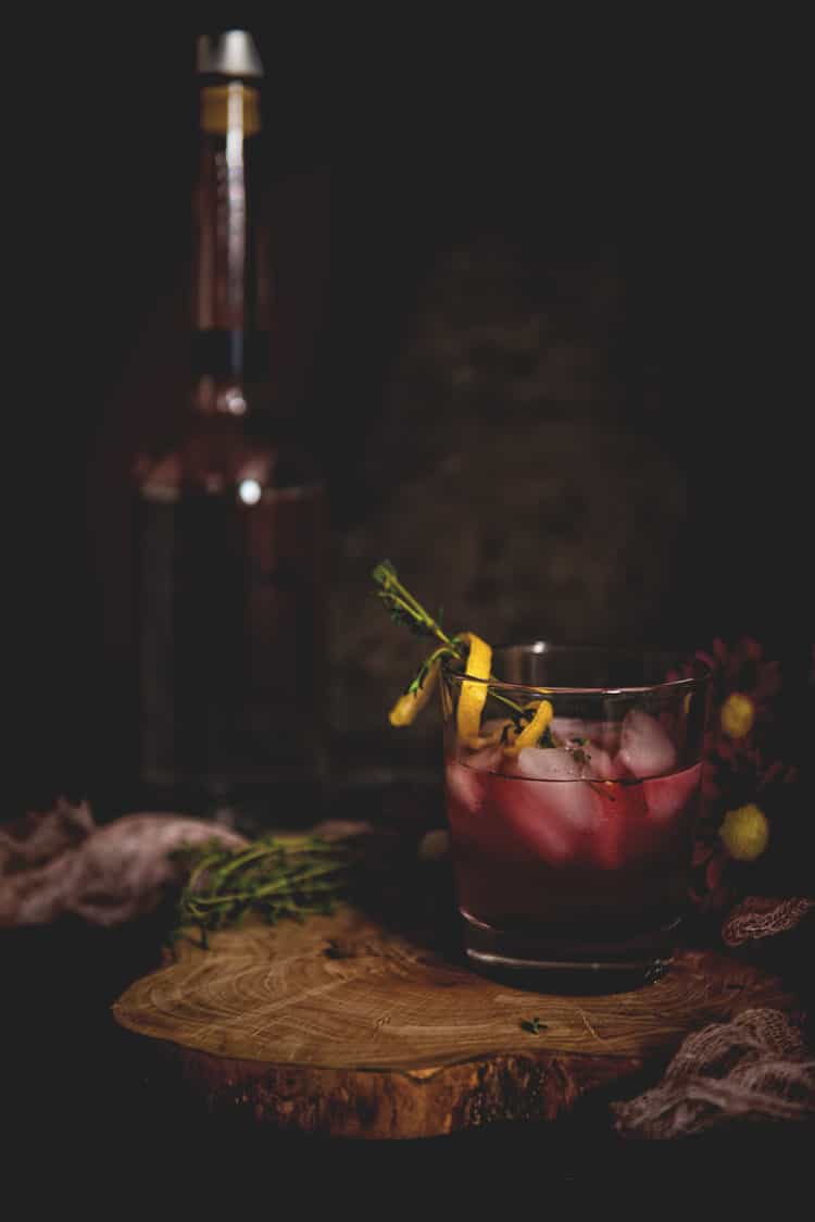 Highball glass with violet huckleberry vodka cocktail, over ice, garnished with lemon twist and thyme sprigs. 
