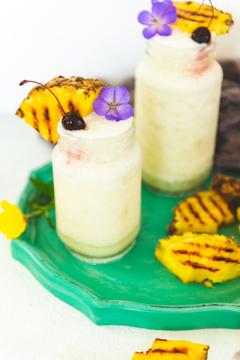 Frozen pina colada cocktails in mason jars garnished with grilled pineapple, cherries and edible flowers. 