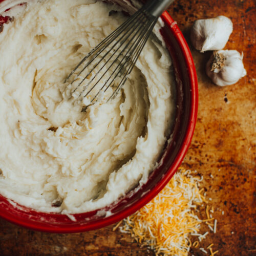 homemade cheesy garlic mashed potatoes in a red bowl with whisk and garlic and cheese on the surface around the bowl