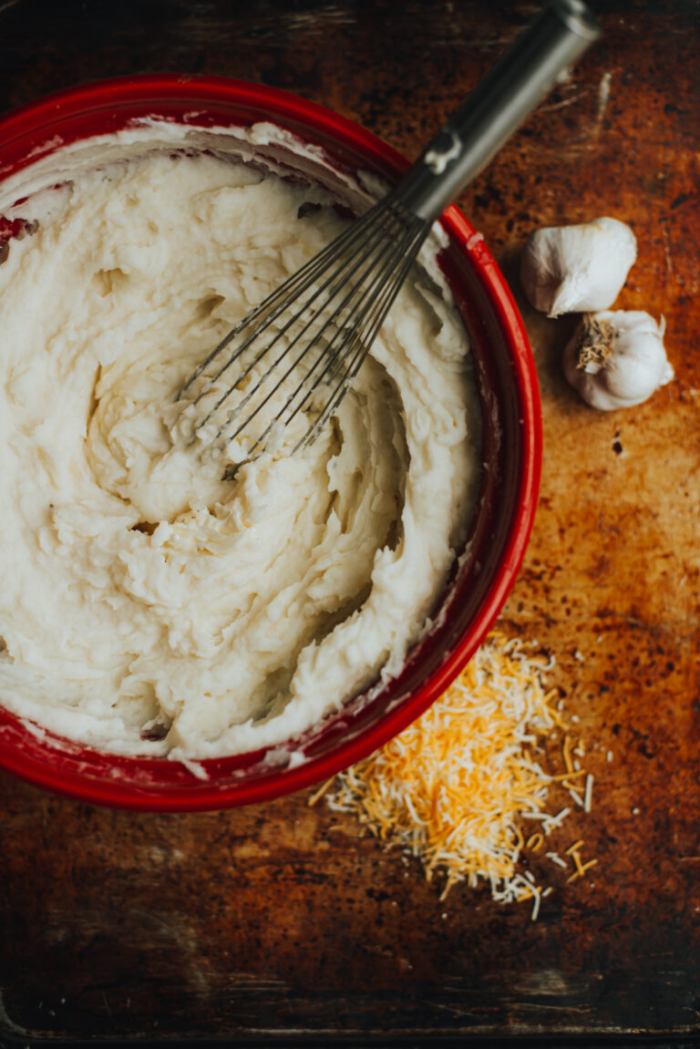 homemade cheesy garlic mashed potatoes in a red bowl with whisk and garlic and cheese on the surface around the bowl