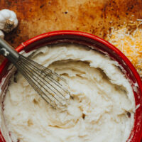 Creamy mashed potatoes in a red bowl with garlic and cheese on the surface around the bowl.