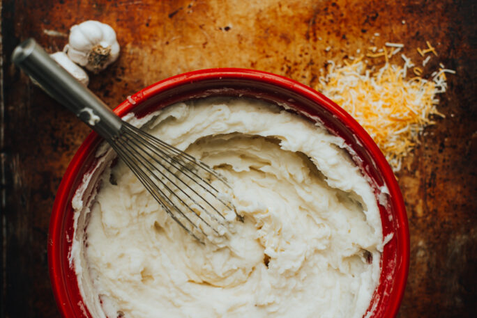 Creamy mashed potatoes in a red bowl with garlic and cheese on the surface around the bowl.