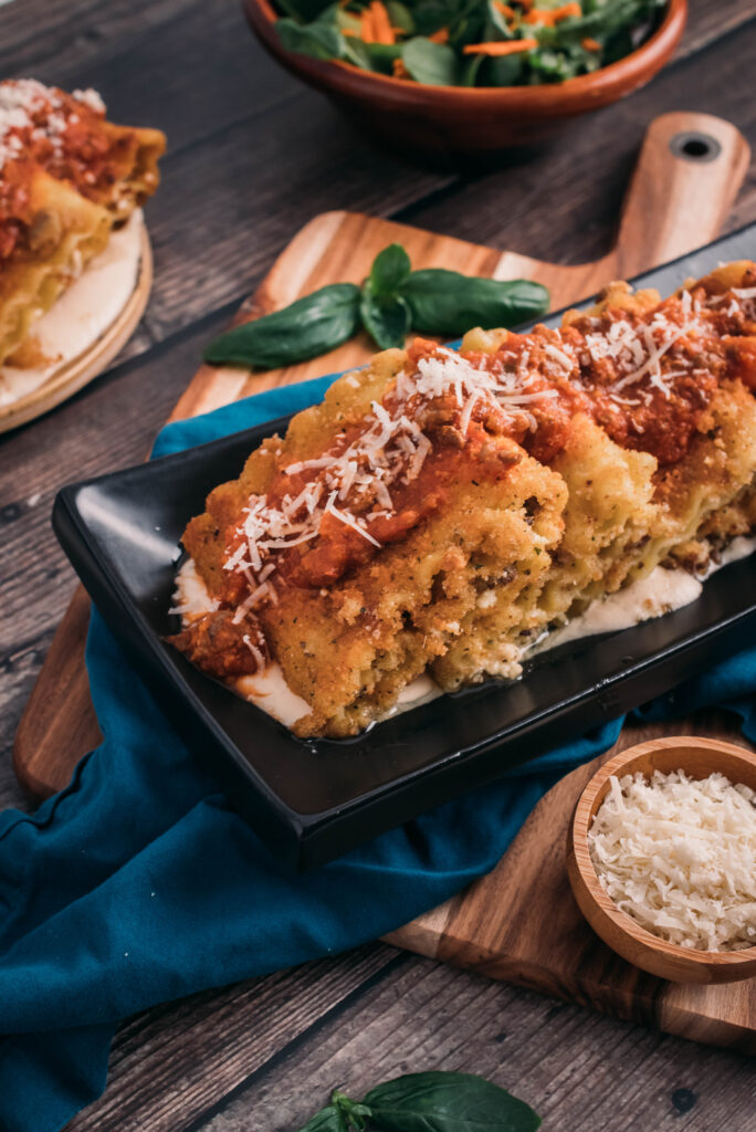 platter of fried lasagne fritta, with alfredo on a black plate, lasagna roll ups and topped with marinara and parmesan.