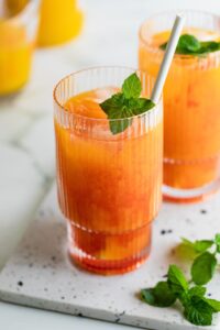 Bright orange red cocktail in a textured glass with a mint sprig and straw.