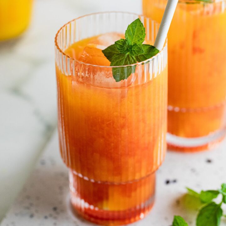 Bright orange red cocktail in a textured glass with a mint sprig and straw.