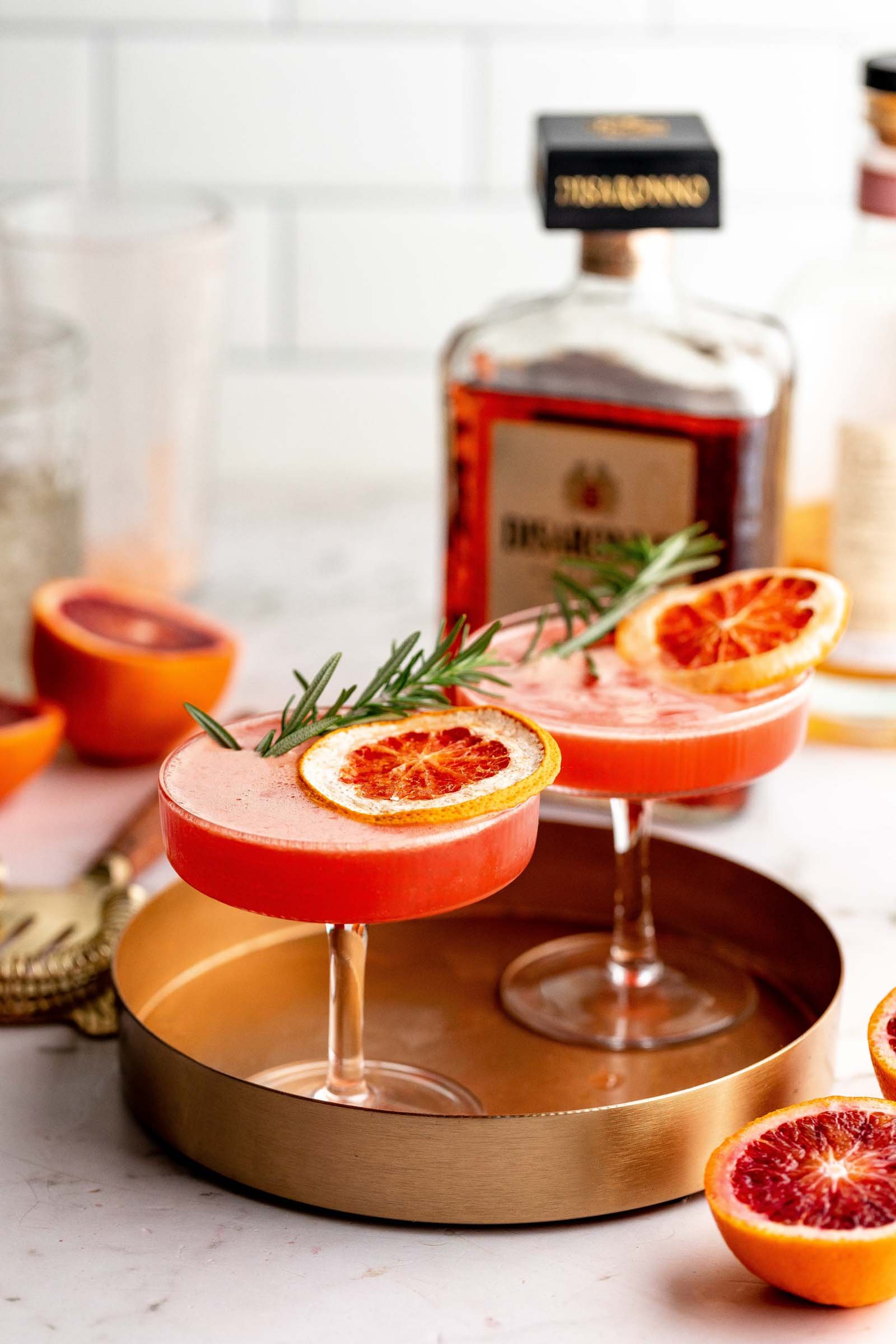 Bright red blood orange cocktails in coupe glasses garnished with rosemary and dried orange wedge on copper platter. 