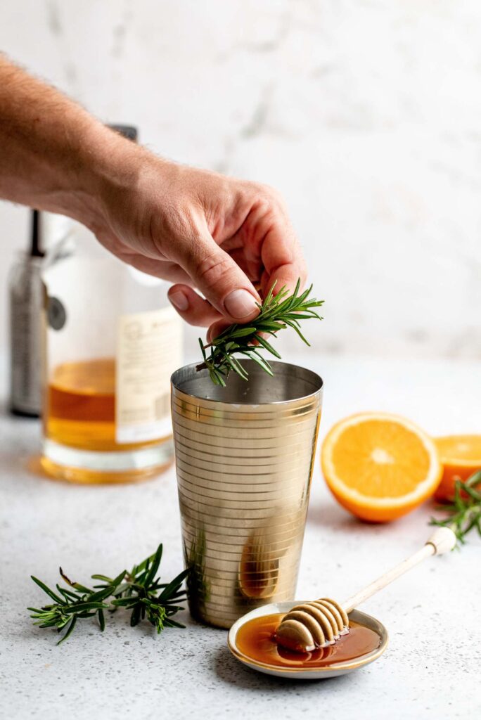 Hand placing rosemary sprig into cocktail shaker.
