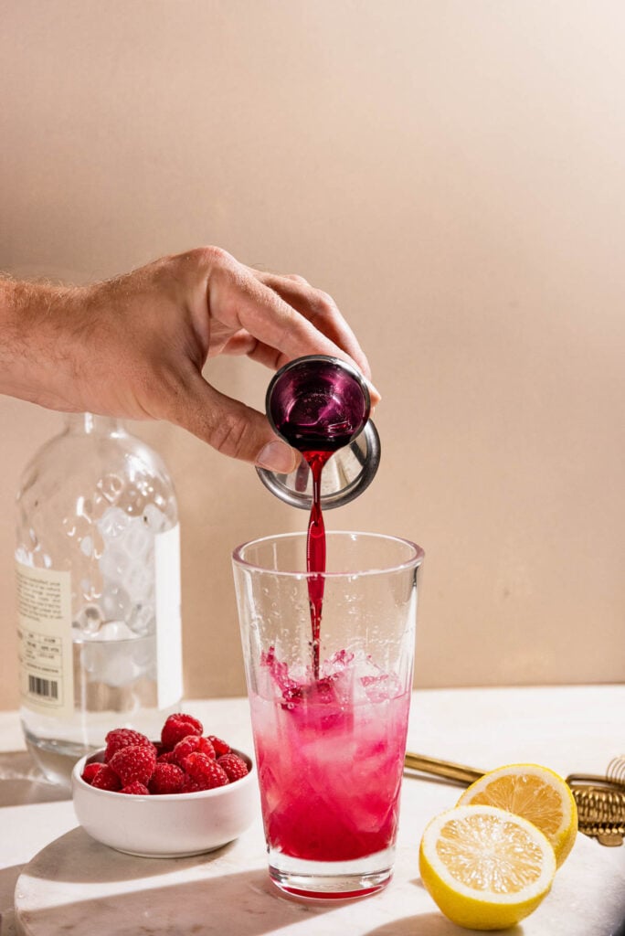 Hand pouring red syrup into a cocktail shaker. 