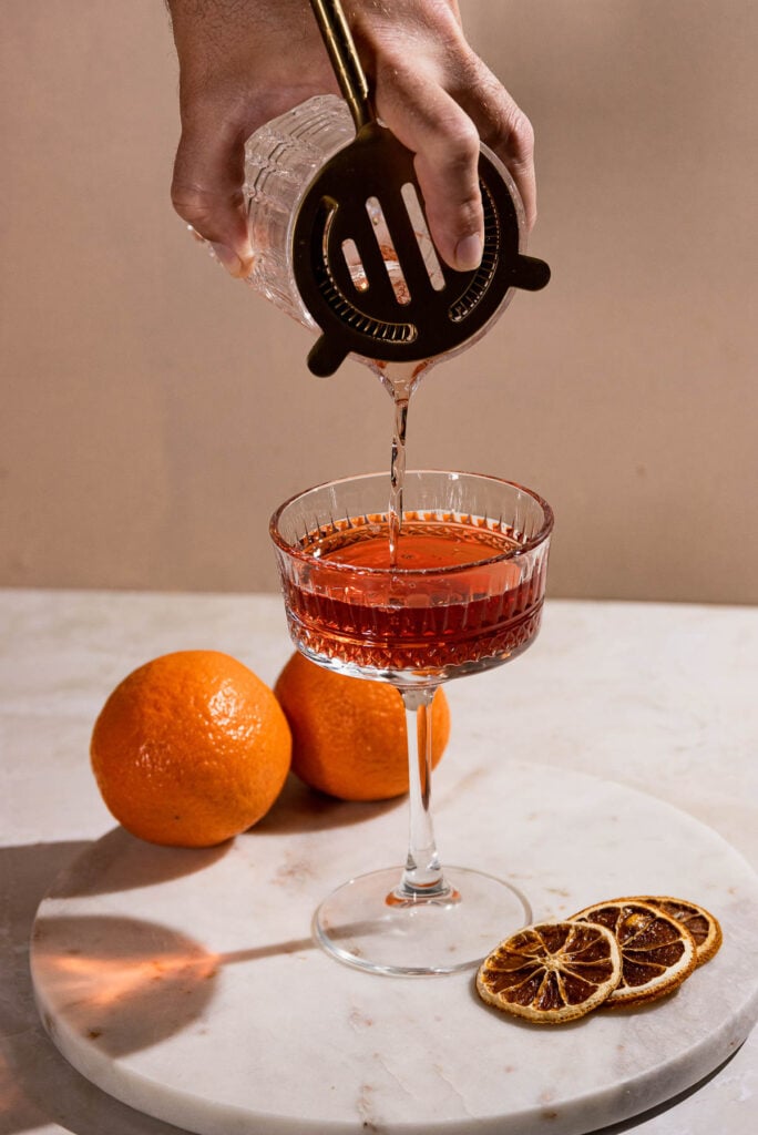 Hand straining cocktail into coupe cocktail glass. 