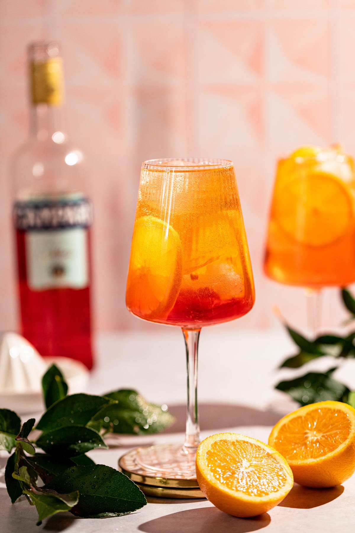 Side shot of a clear glass where you can see the cocktail, glistening oranges and a bottle of Campari in the background. 