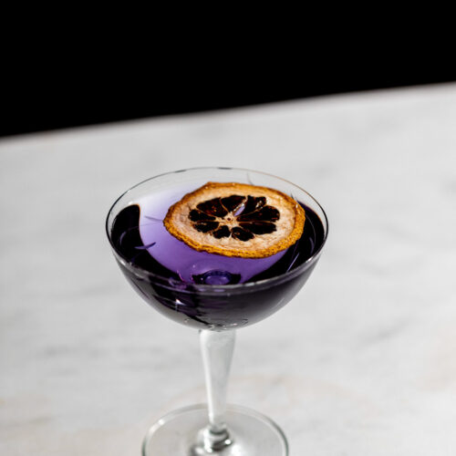 Bright violet aviation cocktail in a coupe glass with a dehydrated lemon.