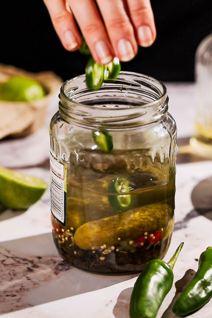 Jalapeno slices being dropped into a jar of pickles.